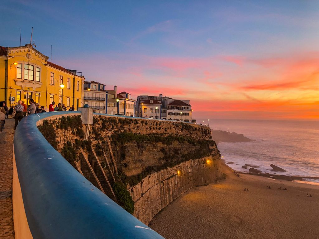 Sunset over Praia dos Pescadores in Ericeira. People stand around a blue and white wall at the top of a cliff overlooking the sand and beach