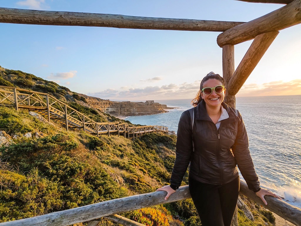 Nicola stands on the wooden staircase leading down to Ribeira d'Ilhas Beach. She is wearing lime green sunglasses and a black jacket and smiling to camera