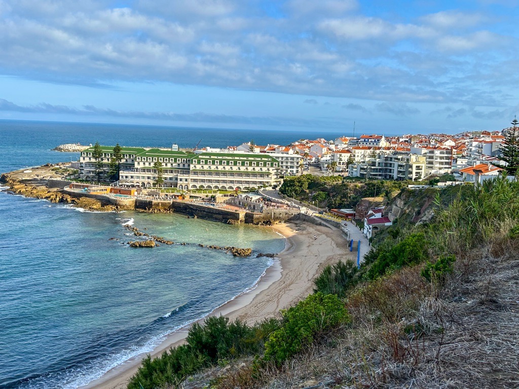The village of Ericeira in the morning. You can see Praia do Sul and Vila Gale, and the water is calm and blue. In the town, most buildings are white with orange roofs