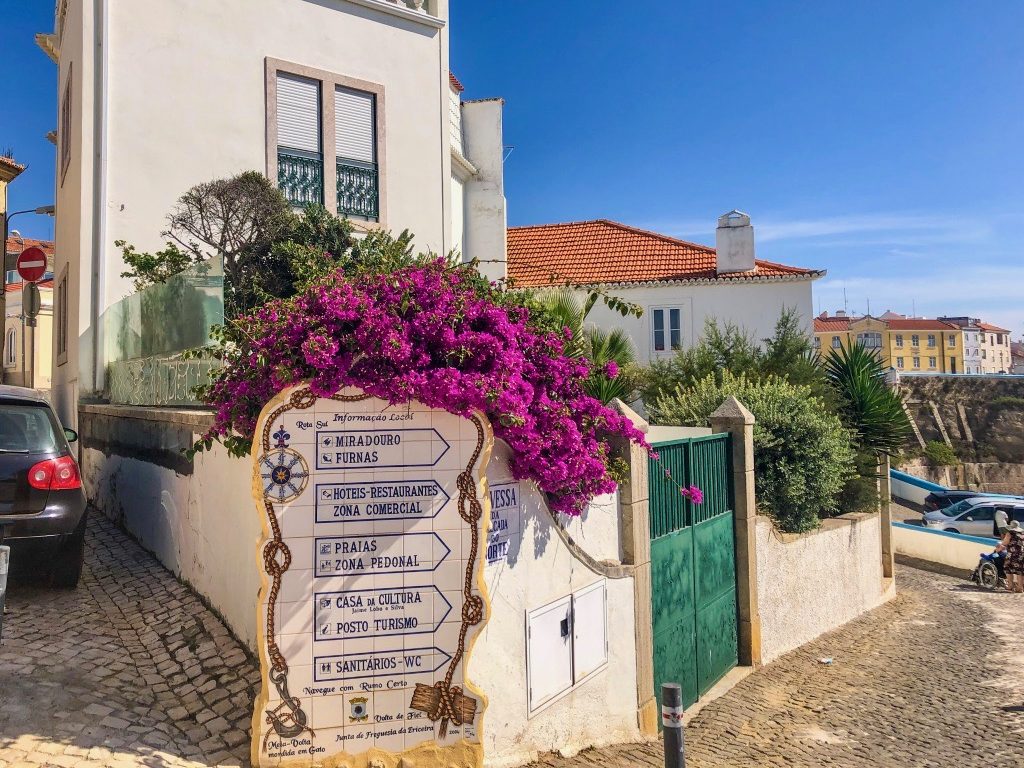 A nautical sign in Ericeira town covered in pink flowers. In the background you can see yellow buildings and the cliffs overlooking the sea