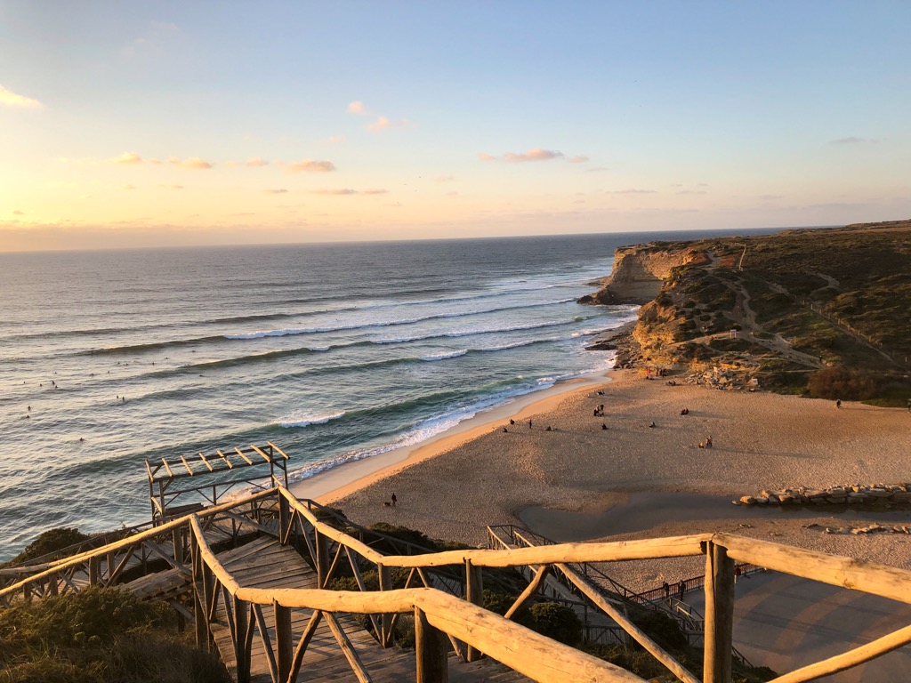 The top of the wooden stairs that lead down to Ribeira d'Ilhas Beach in the late afternoon sun. There are a few surfers in the water and you can see the cliffs in the background
