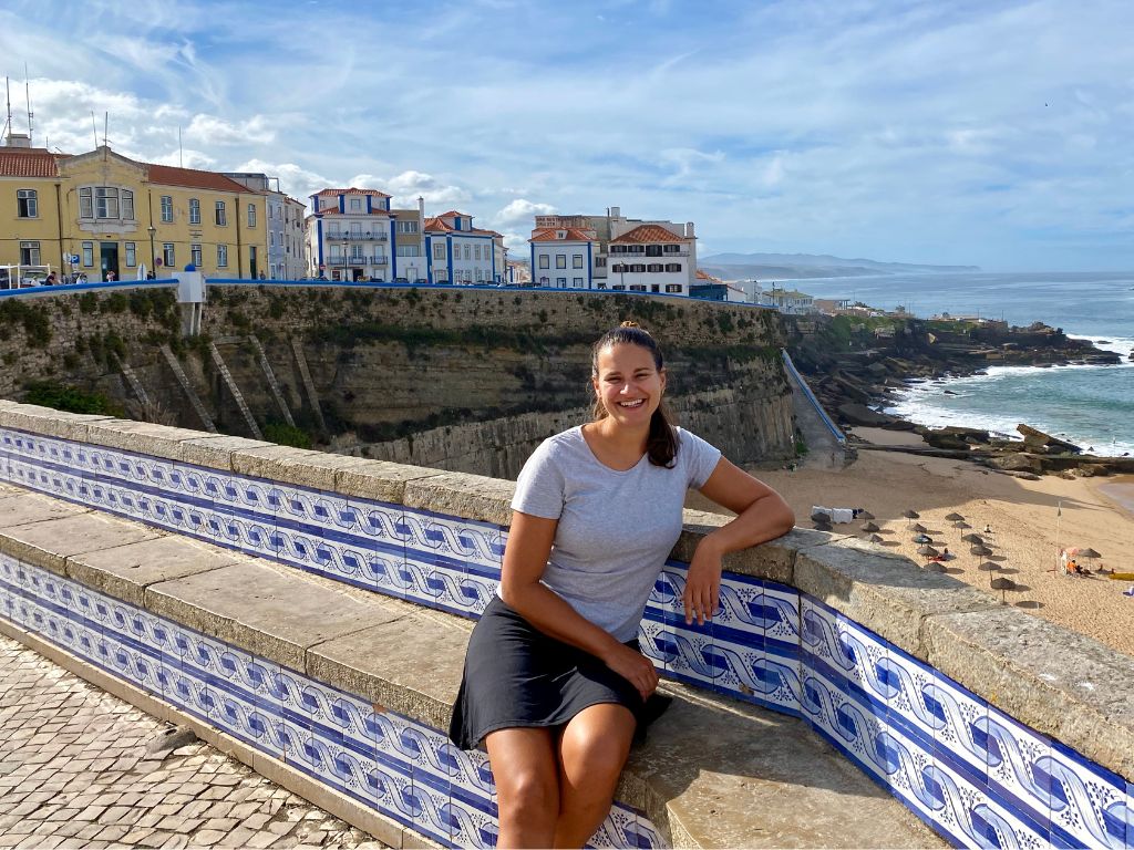 Nicola sits on a tiled bench overlooking the beach in central Ericeira. It's sunny and the buildings in the background are yellow and white with blue trim.